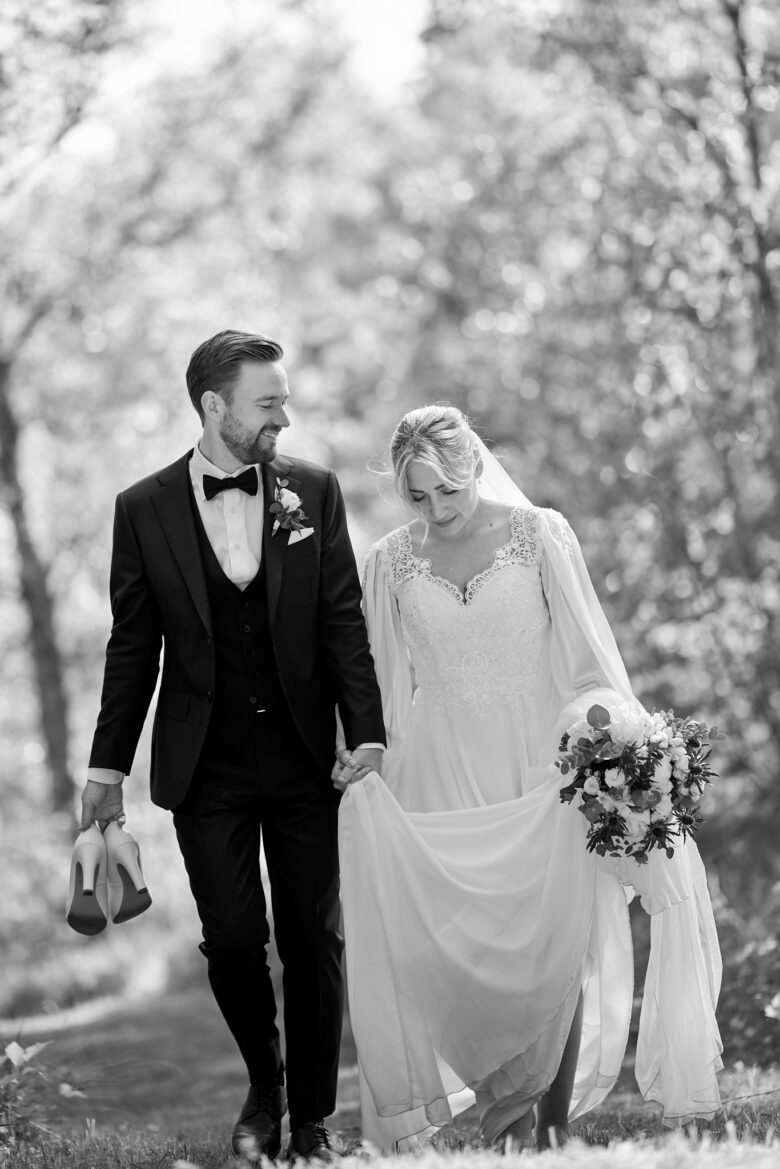 Bröllop-på Balingsholm herrgård-svartvit-bild-på-barfota-brud-med-bukett-och brudgum--som-går-brevis-och-håller-hennes-skor